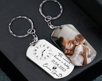 May include: Silver keychain with two dog tags. One tag has a clock face and the text "The moment I became a daddy 01.04.2023 Emma Grace". The other tag has a photo of a father and baby.