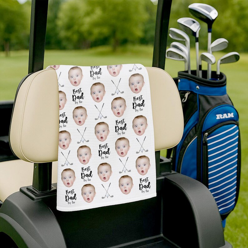 May include: A white golf towel with a repeating pattern of a baby's face and the text "Best Dad By Far". The towel is draped over a golf cart seat, with golf clubs in a blue and black bag in the background.