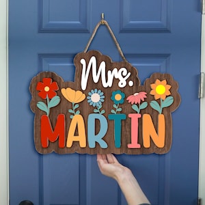 May include: A wooden door hanger with the text "Mrs. Martin" in colorful letters. The letters are surrounded by colorful flowers with green stems.