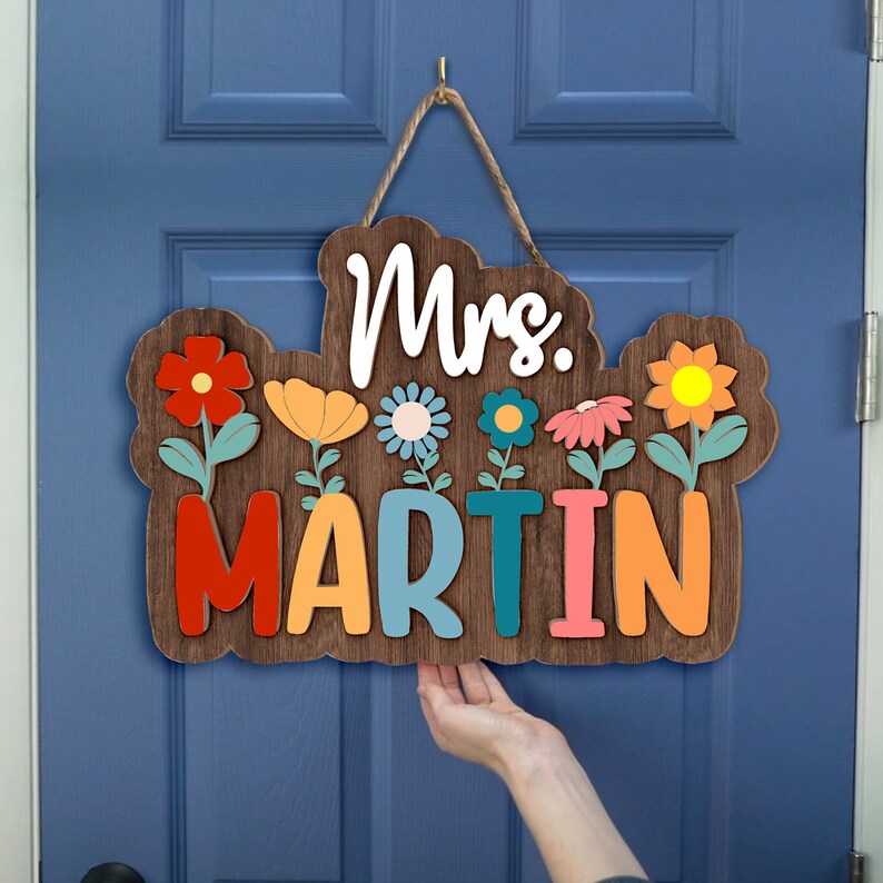 May include: A wooden door hanger with the text "Mrs. Martin" in colorful letters. The letters are surrounded by colorful flowers with green stems.