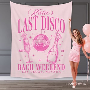 May include: Pink bachelorette party backdrop with the text "Katie's Last Disco" and "Bach Weekend Las Vegas, Nevada". The backdrop also features a disco ball, champagne bottle, and champagne flute.