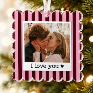 May include: A square scalloped Christmas ornament featuring a pink and dark red vertical striped border. The center displays a photograph of two people embracing, with the text I love you and a heart symbol printed in black below the image.