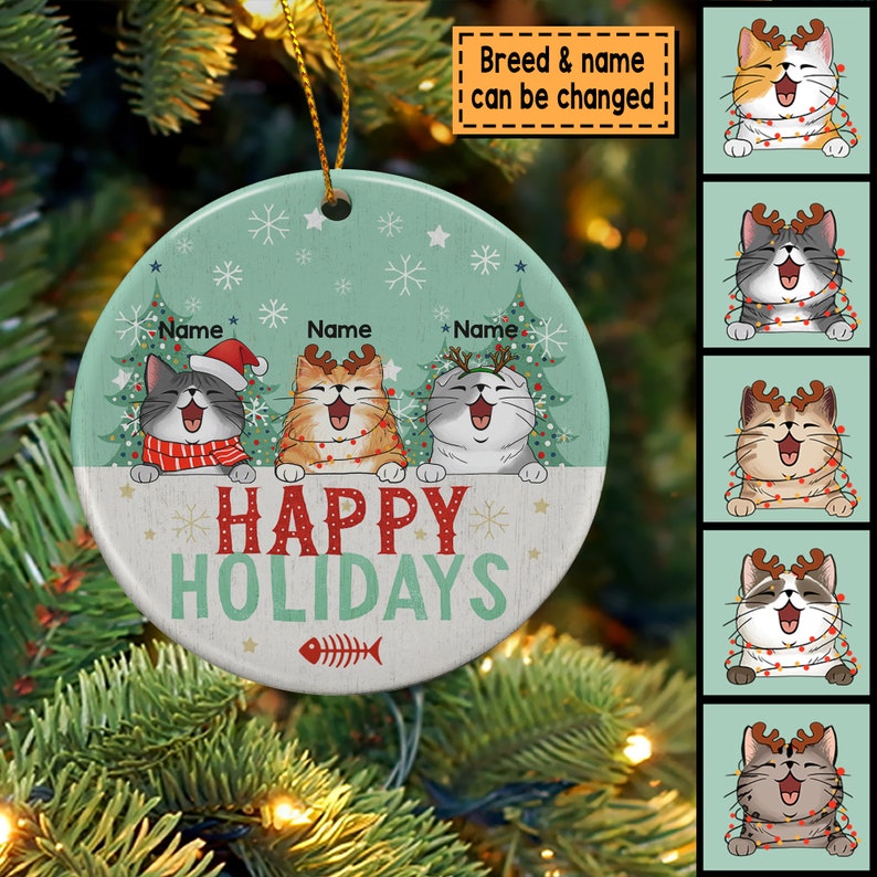 May include: A round, white ceramic ornament with a green background and three cartoon cats wearing reindeer antlers. The cats are surrounded by snowflakes and Christmas trees. The text "Happy Holidays" is written in red letters on the ornament.