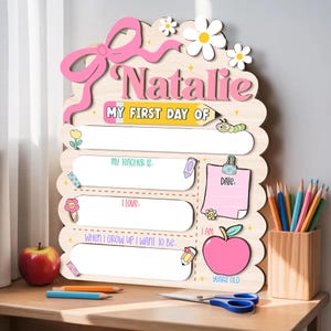 May include: A wooden sign for the first day of school, with the name "Natalie" in pink. The sign has spaces to write details about the day, decorated with flowers, a bow, and a pink apple. A pencil holder with coloured pencils is visible.