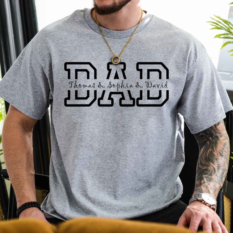 K&ouml;nnte beinhalten: Graues T-Shirt mit einer schwarzen Grafik, auf der "DAD" steht, darunter die Namen "Thomas & Sophia & David".