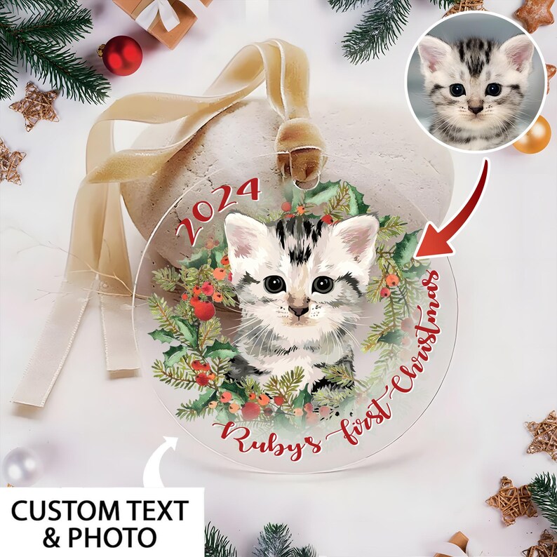 May include: A clear, round Christmas ornament featuring a watercolor illustration of a kitten within a wreath of greenery and berries. The ornament has the text "2024" and "Ruby's first Christmas" in red script. A ribbon is attached for hanging.