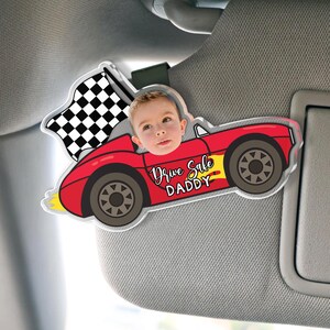 May include: A red car-shaped sun visor accessory with a child's face in the driver's seat. The car has black wheels, a checkered flag, and the text "Drive Safe DADDY". The accessory is attached to a car visor.