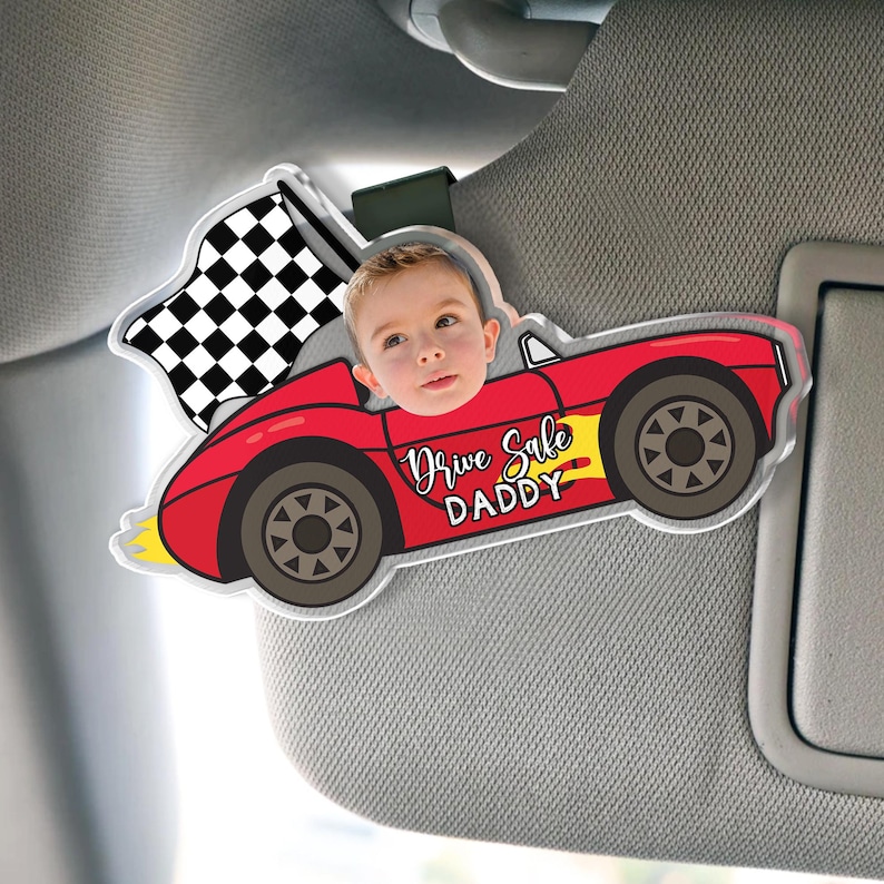May include: A red car-shaped sun visor accessory with a child's face in the driver's seat. The car has black wheels, a checkered flag, and the text "Drive Safe DADDY". The accessory is attached to a car visor.