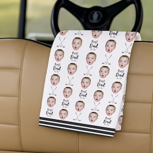 May include: White golf towel featuring a repeating pattern of a baby's face, golf clubs, and the text "Best Dad By far." The towel has black stripes at the bottom and is folded over a golf cart seat.
