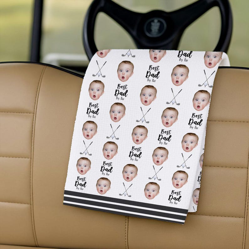 May include: White golf towel featuring a repeating pattern of a baby's face, golf clubs, and the text "Best Dad By far." The towel has black stripes at the bottom and is folded over a golf cart seat.