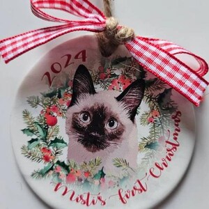 May include: A round Christmas ornament featuring a Siamese cat portrait surrounded by a festive wreath. The ornament has the year "2024" and the text "Misti's first Christmas" in red script. It is adorned with a red and white checkered ribbon.