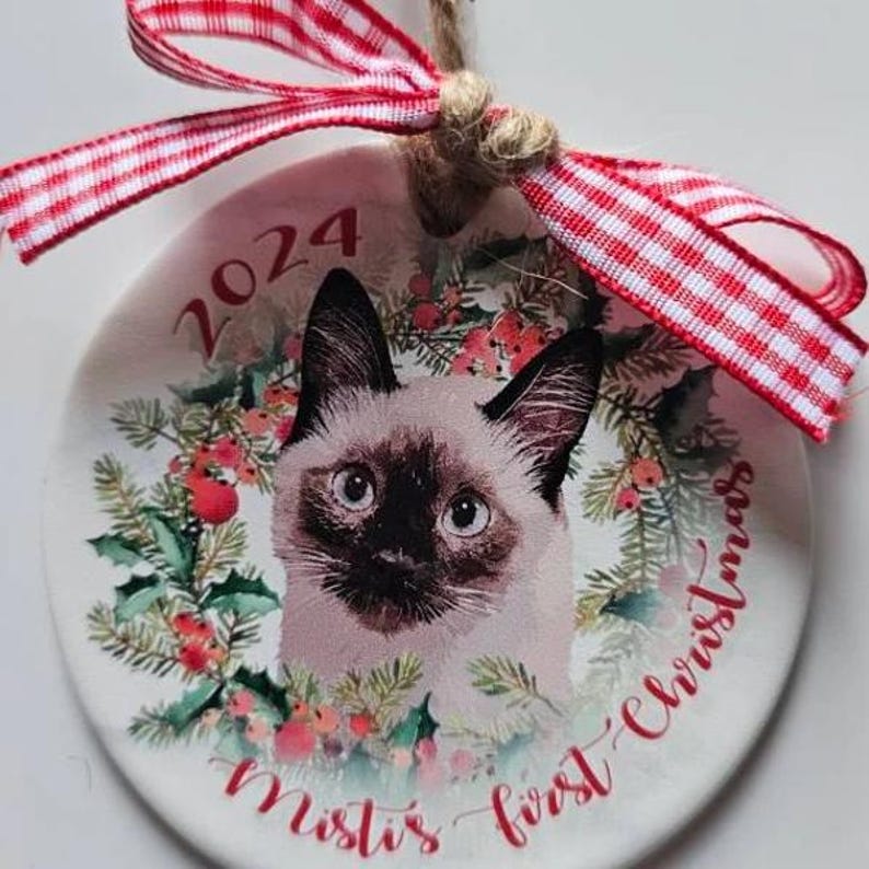 May include: A round Christmas ornament featuring a Siamese cat portrait surrounded by a festive wreath. The ornament has the year "2024" and the text "Misti's first Christmas" in red script. It is adorned with a red and white checkered ribbon.