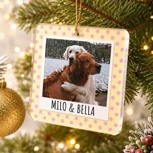 May include: A square acrylic Christmas ornament featuring a light yellow background with pink polka dots. The center displays a photograph of two dogs hugging by a lake, with the text MILO & BELLA printed in black below the image.