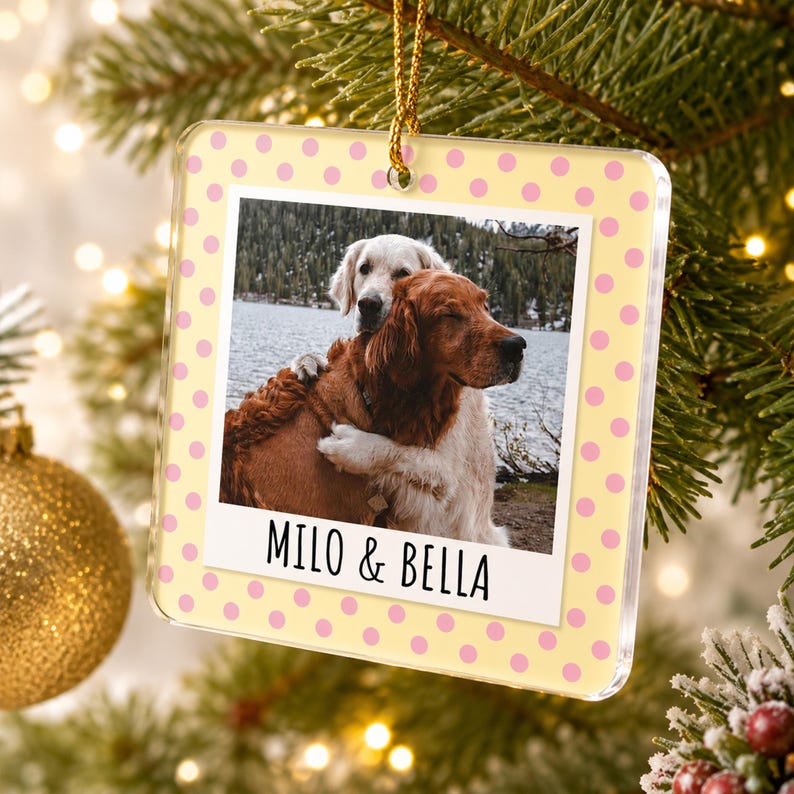 May include: A square acrylic Christmas ornament featuring a light yellow background with pink polka dots. The center displays a photograph of two dogs hugging by a lake, with the text MILO & BELLA printed in black below the image.