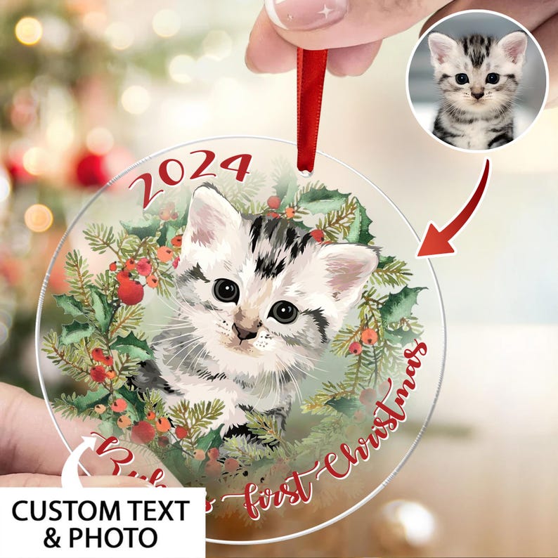 May include: Clear, round Christmas ornament featuring a gray and white kitten surrounded by a holly wreath and red berries. The ornament has the text "2024" and "Baby's first Christmas" in red script. Includes a red ribbon for hanging.