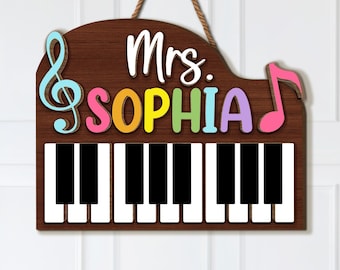 May include: Wooden piano-shaped sign with a brown background, a piano keyboard, and the name "Mrs. SOPHIA" in colorful letters. A treble clef and a musical note are on the left and right sides. The sign hangs from a brown rope.