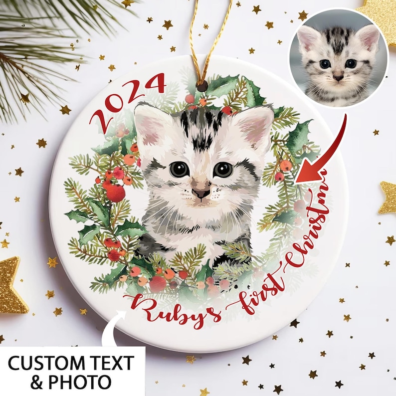 May include: A white ceramic Christmas ornament featuring a gray and white kitten surrounded by a wreath of holly and berries. The ornament has the text "2024" and "Ruby's first Christmas" in red script. A small photo of the kitten is in the upper right.