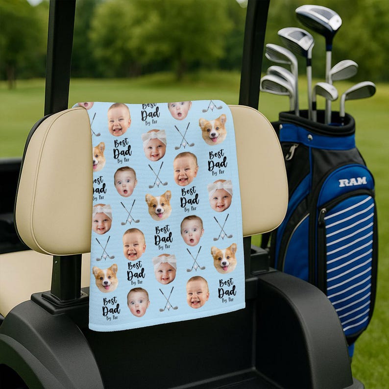May include: A light blue golf towel with a repeating pattern of baby faces, corgis, and golf clubs. The text "Best Dad by far" is printed on the towel. The towel is placed on a golf cart.