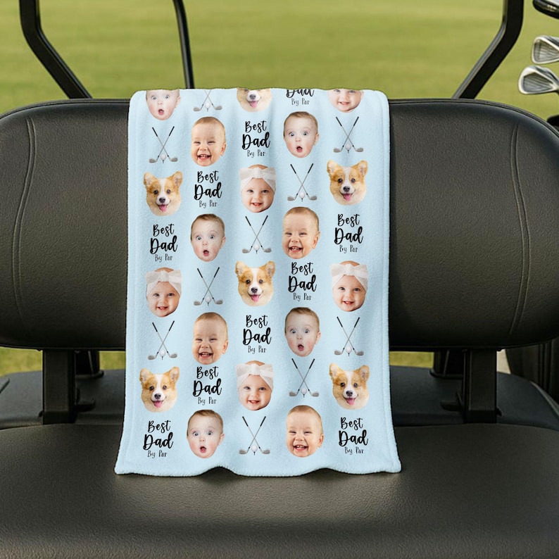May include: A light blue golf towel with a repeating pattern of baby faces, corgi faces, and crossed golf clubs. The words "Best Dad By Far" are printed in black script. This towel is a potential Father's Day gift.
