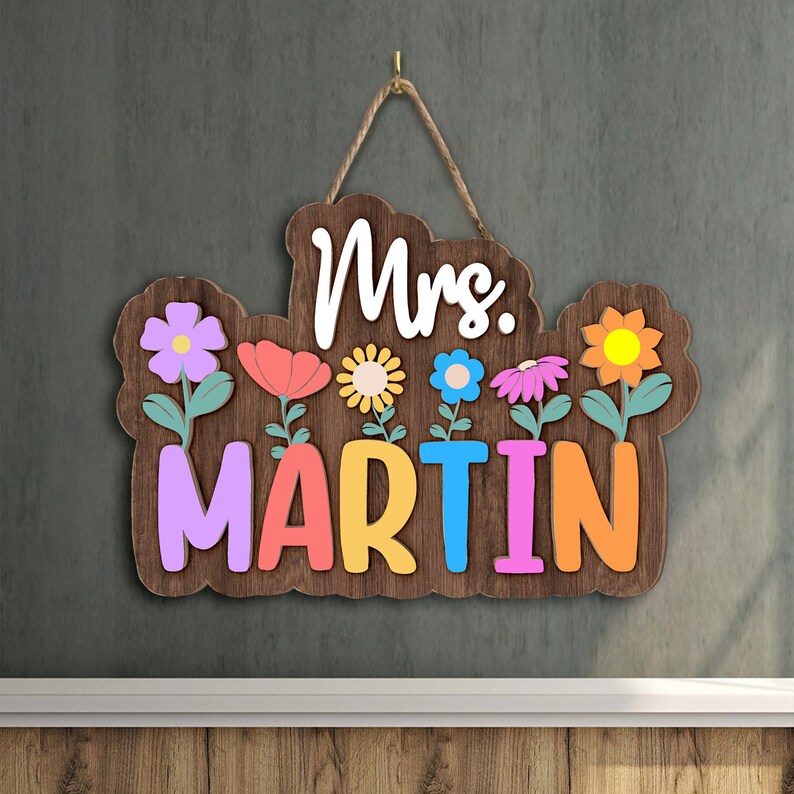May include: A wooden sign with the text "Mrs. MARTIN" in colorful letters. The sign is decorated with flowers in various colors, including pink, yellow, orange, and blue.