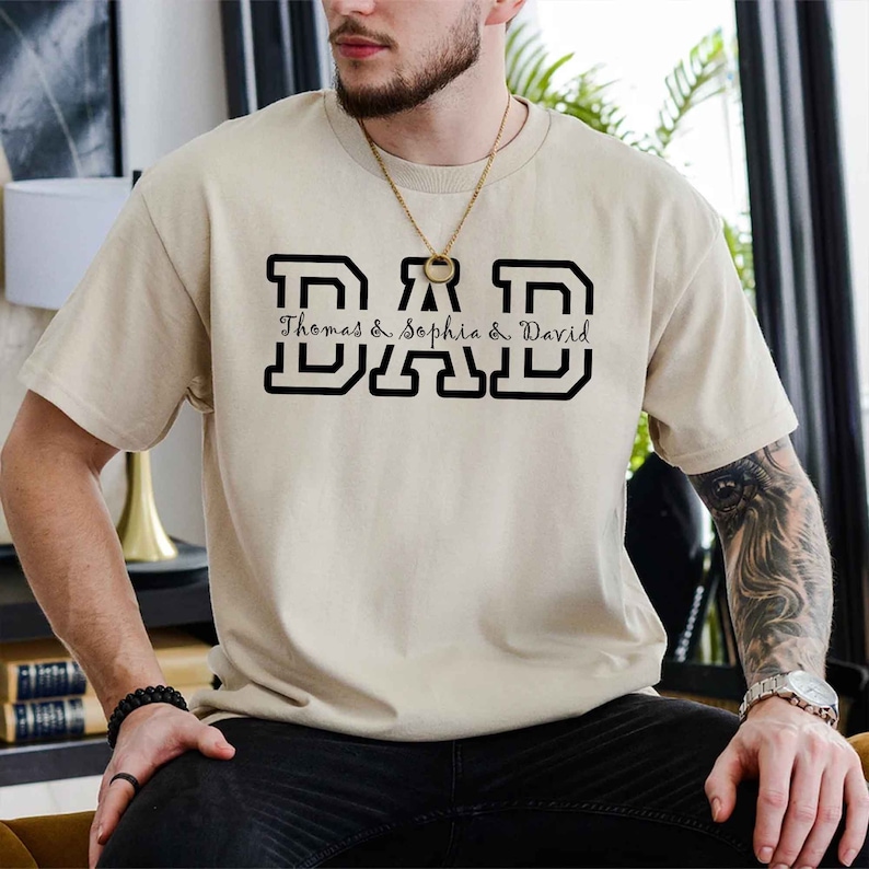 K&ouml;nnte beinhalten: Ein beiges T-Shirt mit einer schwarzen Grafik, auf der "DAD" in gro&szlig;en Blockbuchstaben steht. Unter den Buchstaben steht "Thomas & Sophia & David" in einer kleineren, kursiv geschriebenen Schrift.