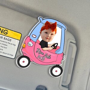 May include: A pink and blue car-shaped car window decal with a photo of a baby wearing a red bow. The decal reads "Drive Safe Dad". The decal is attached to a car visor.