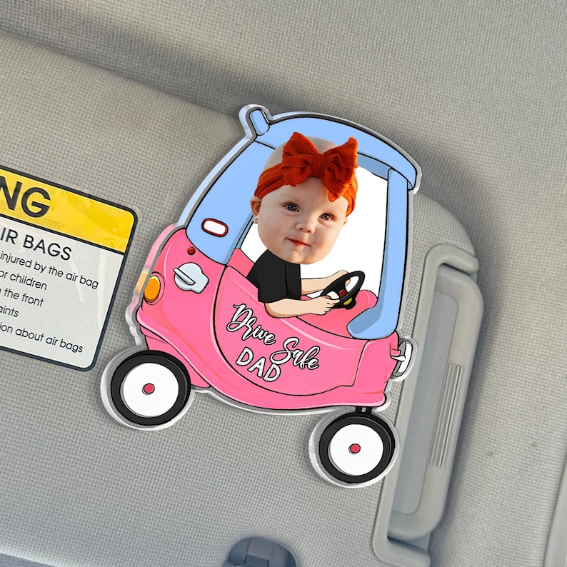 May include: A pink and blue car-shaped car window decal with a photo of a baby wearing a red bow. The decal reads "Drive Safe Dad". The decal is attached to a car visor.