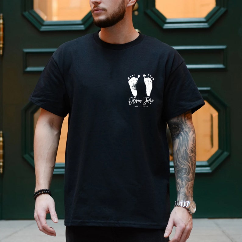May include: Black t-shirt with a white design of two baby footprints and the text "Olivia June" and "April 11, 2024".