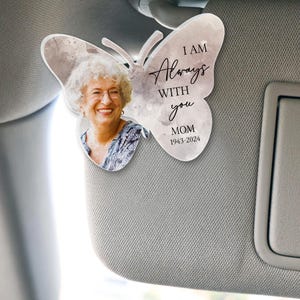 May include: A white car sun visor with a gray and white butterfly-shaped memorial ornament. The ornament features a photo of a woman and the text "I AM Always WITH you MOM 1943-2024".