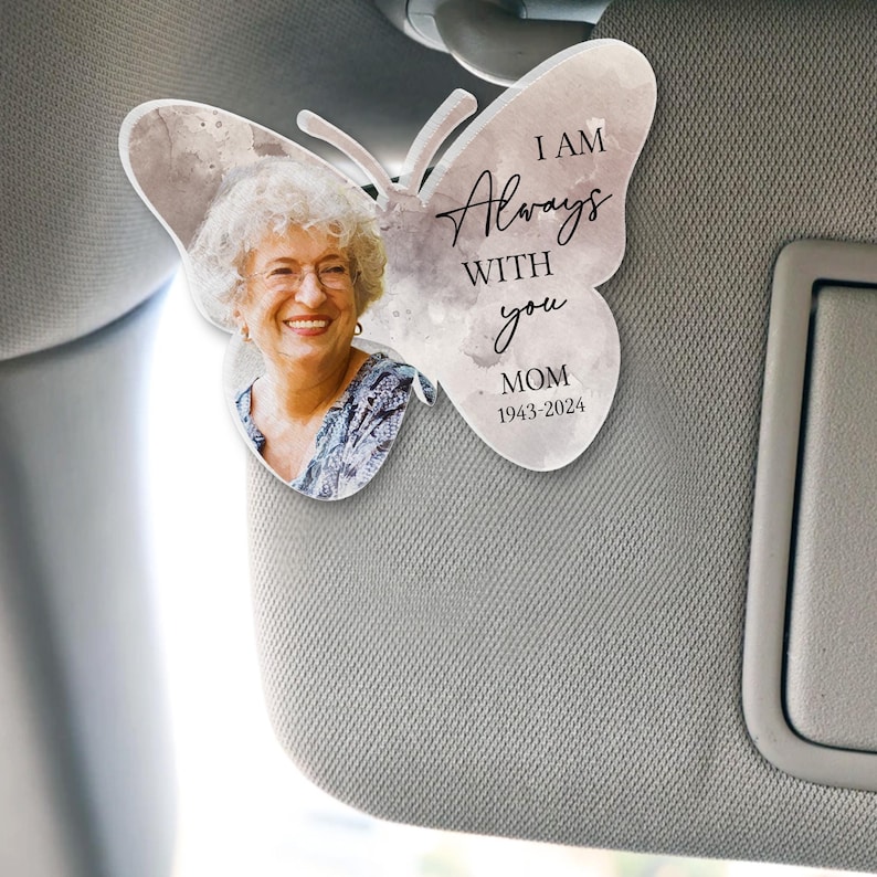 May include: A white car sun visor with a gray and white butterfly-shaped memorial ornament. The ornament features a photo of a woman and the text "I AM Always WITH you MOM 1943-2024".