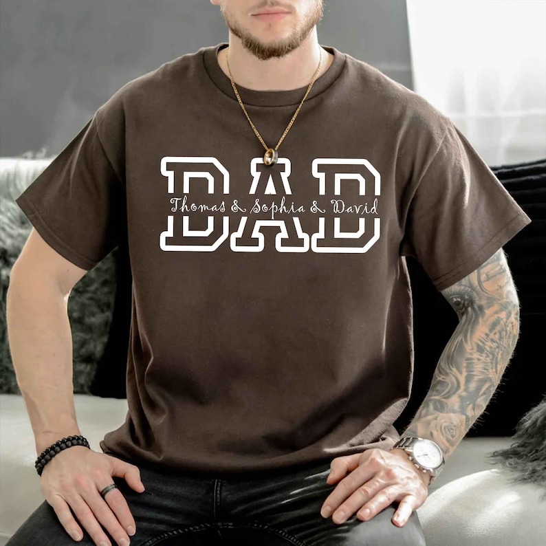 K&ouml;nnte beinhalten: Braunes T-Shirt mit wei&szlig;em Text, der "DAD Thomas & Sophia & David" in einer Schriftart im College-Stil lautet.