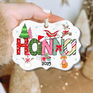 May include: A personalized Christmas ornament with the name "HANNA" in colorful, festive lettering. The ornament features a snowman, reindeer, sleigh, and fox, with the year "2025" displayed. The ornament is held by a hand.