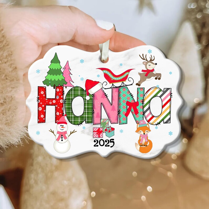 May include: A personalized Christmas ornament with the name "HANNA" in colorful, festive lettering. The ornament features a snowman, reindeer, sleigh, and fox, with the year "2025" displayed. The ornament is held by a hand.