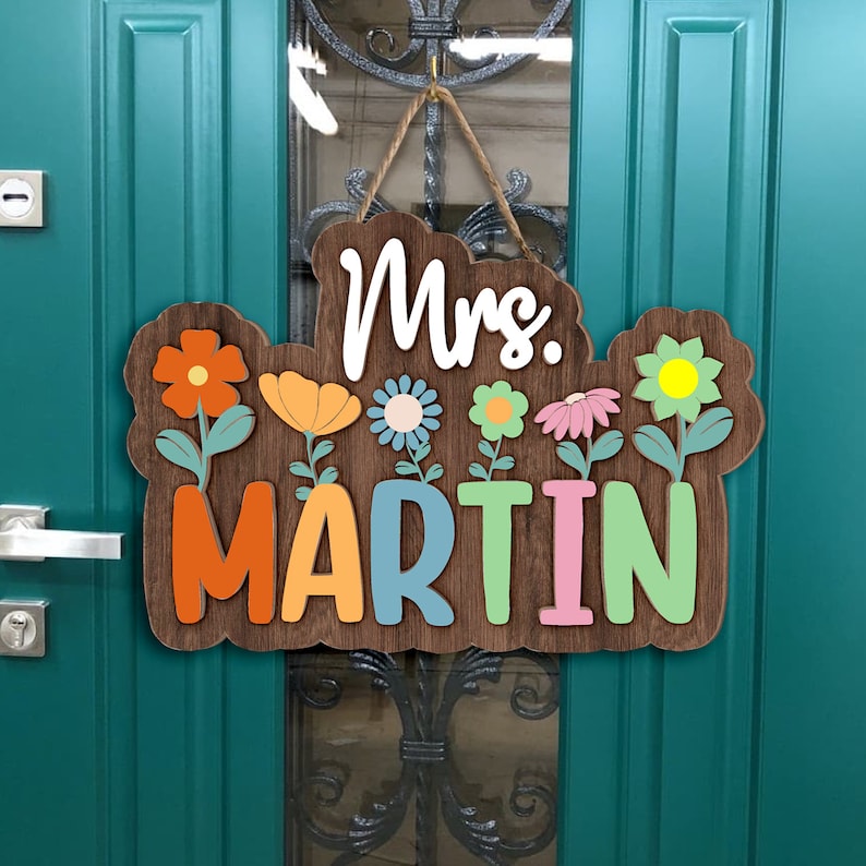 May include: A wooden door hanger with the text "Mrs. Martin" in colorful letters. The letters are surrounded by colorful flowers with green stems.
