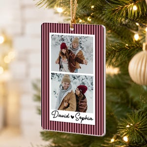 May include: A rectangular hanging ornament featuring a dark pink and black vertical striped border. The center displays two stacked photographs of a couple in a snowy landscape, with the text David and Sophia printed below.