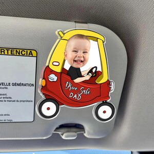 May include: A red and yellow car-shaped visor clip featuring a smiling baby. The clip has the words "Drive Safe DAD" in a cursive font. The clip is attached to a car visor.