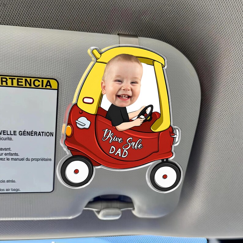 May include: A red and yellow car-shaped visor clip featuring a smiling baby. The clip has the words "Drive Safe DAD" in a cursive font. The clip is attached to a car visor.
