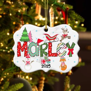 May include: A white ceramic Christmas ornament with the name "MORLEY" in colorful letters. The ornament features festive illustrations of a Christmas tree, snowman, reindeer, fox, and a sleigh. The year "2025" is also displayed.
