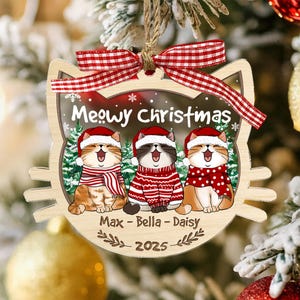 May include: Wooden Christmas ornament shaped like a cat's face, featuring three cartoon cats wearing Santa hats and scarves. The ornament has the text "Meowy Christmas" and the names "Max - Bella - Daisy" with the year 2025.