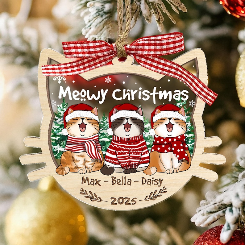 May include: Wooden Christmas ornament shaped like a cat's face, featuring three cartoon cats wearing Santa hats and scarves. The ornament has the text "Meowy Christmas" and the names "Max - Bella - Daisy" with the year 2025.