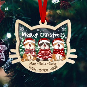 May include: Wooden Christmas ornament shaped like a cat's head, featuring three cartoon cats wearing Santa hats and scarves. The ornament says "Meowy Christmas" and includes the names "Max - Bella - Daisy" and the year 2024.