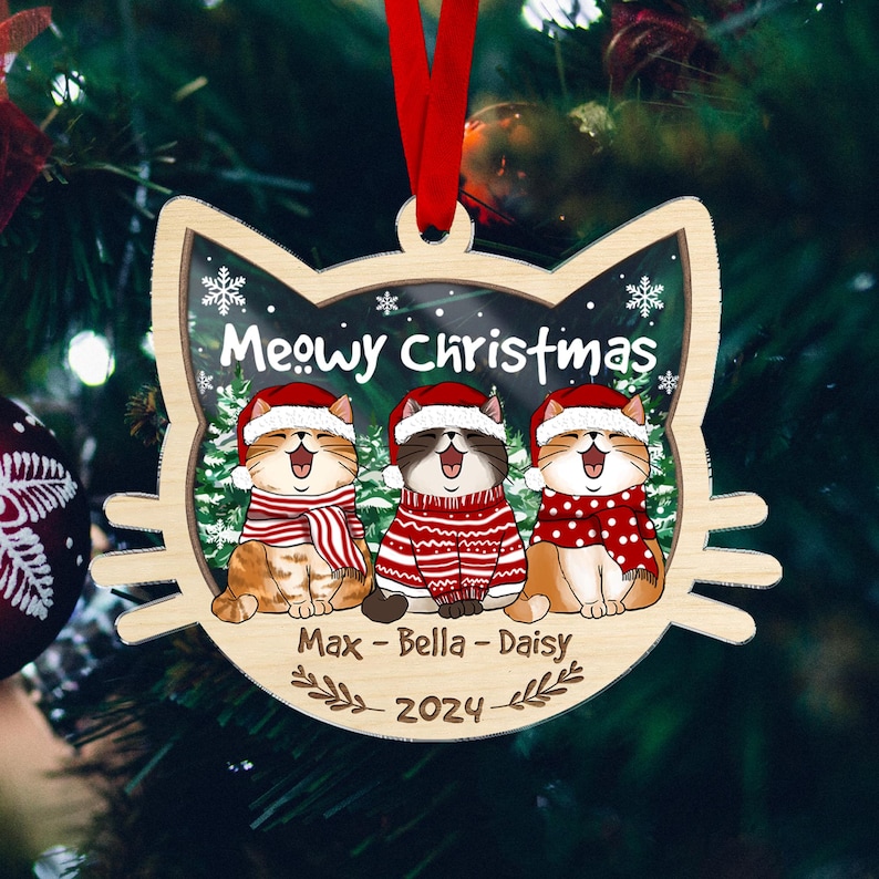 May include: Wooden Christmas ornament shaped like a cat's head, featuring three cartoon cats wearing Santa hats and scarves. The ornament says "Meowy Christmas" and includes the names "Max - Bella - Daisy" and the year 2024.