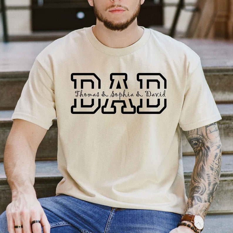 K&ouml;nnte beinhalten: Ein beiges T-Shirt mit einer schwarzen Grafik, auf der "DAD" in gro&szlig;en Blockbuchstaben steht. Unter den Buchstaben steht "Thomas & Sophia & David" in einer kleineren, kursiv geschriebenen Schrift.
