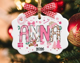 Könnte beinhalten: Ein weißer, geschwungener Keramik-Weihnachtsanhänger an einer Jutekordel mit rot-weiß karierter Schleife. Der Anhänger zeigt den Namen ANNA in rosa und leopardenmusterfarbenen Buchstaben, umgeben von Rentieren, rosa Bäumen und der Jahreszahl 2026.