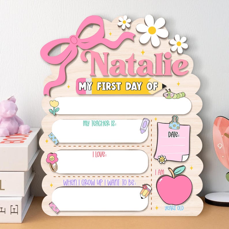 May include: A wooden sign for the first day of school, with the name "Natalie" in pink. The sign has spaces to write details about the day, decorated with a bow, flowers, and a pencil. Includes the text "MY FIRST DAY OF".