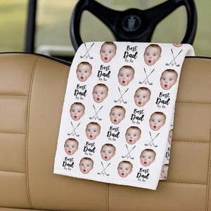 May include: White golf towel with a repeating pattern of a baby's face, golf clubs, and the text "Best Dad By Far." The towel is draped over a golf cart's armrest, with the steering wheel in the background.