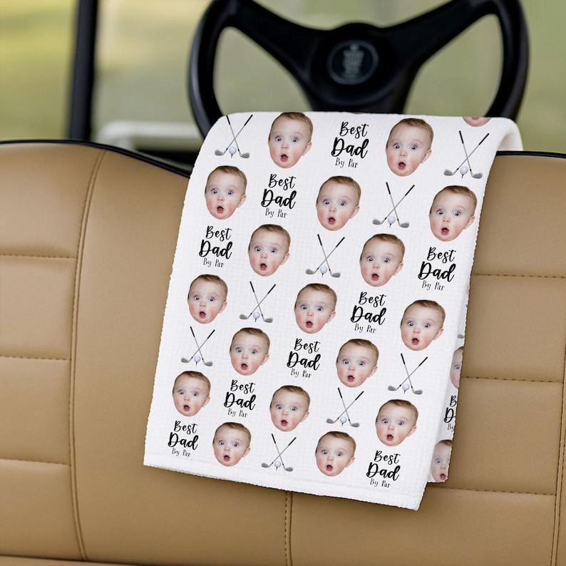 May include: White golf towel with a repeating pattern of a baby's face, golf clubs, and the text "Best Dad By Far." The towel is draped over a golf cart's armrest, with the steering wheel in the background.
