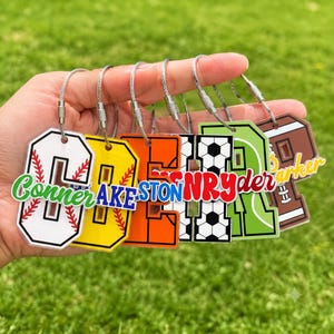 May include: A set of six acrylic letter keychains with metal wire loops. Each letter features a unique sports-themed design including baseball, soccer, and football patterns, with personalized names printed in various colors and fonts across them.