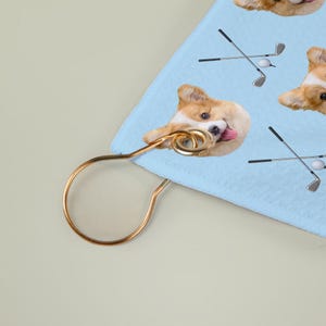 May include: A light blue golf towel with a repeating pattern of a corgi dog, golf clubs, and golf balls. The towel has a gold-coloured metal hanger attached.