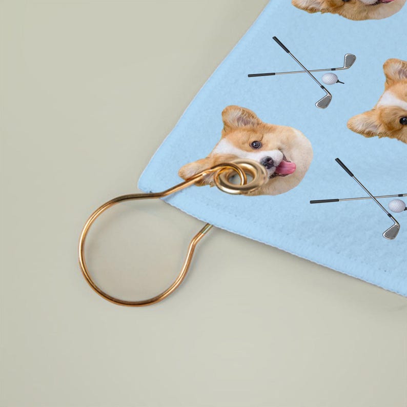 May include: A light blue golf towel with a repeating pattern of a corgi dog, golf clubs, and golf balls. The towel has a gold-coloured metal hanger attached.